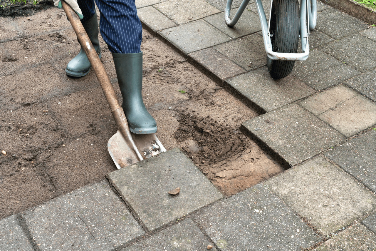 Persoon met laarzen aan haalt met een schop tegels uit een tuin.