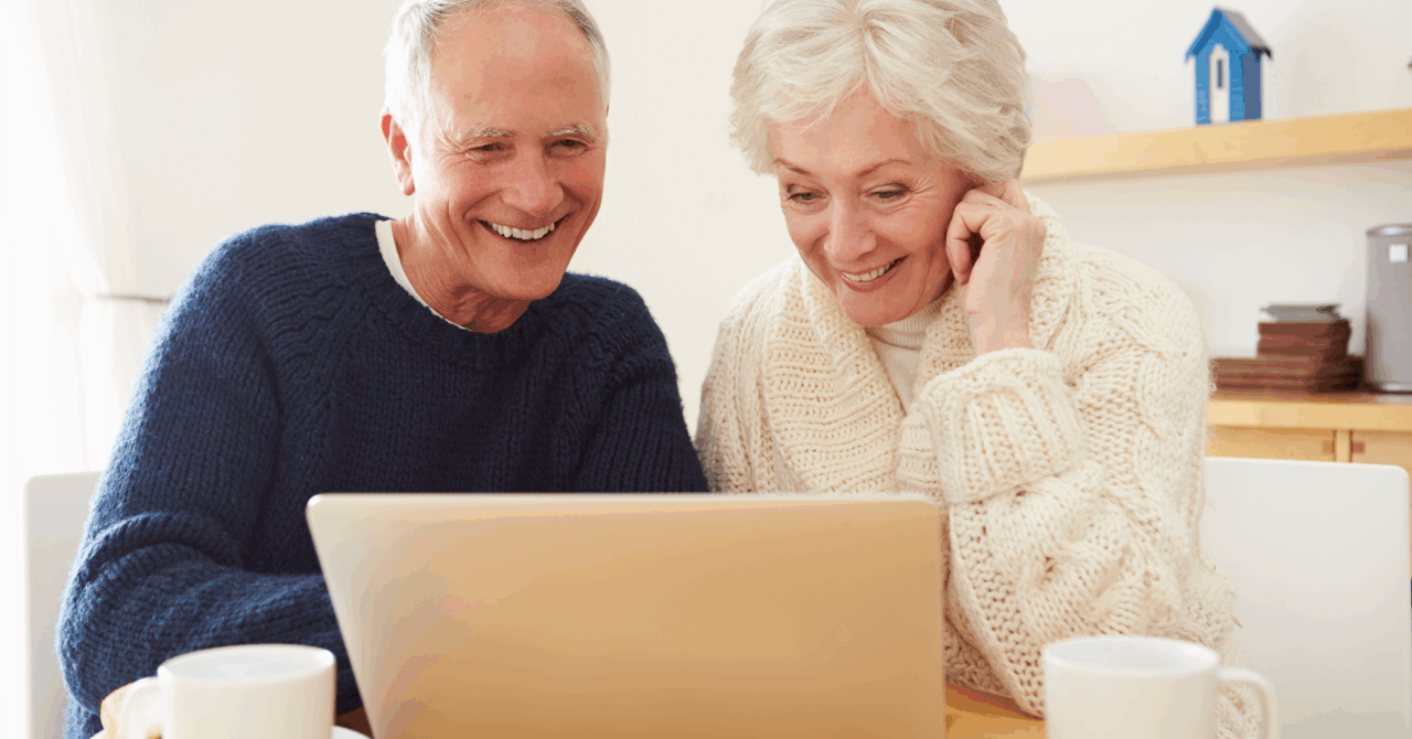Oudere man en vrouw met grijs haar en warme wintertruien kijken naar een laptopscherm en lachen